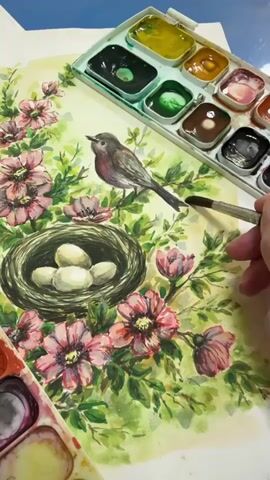 The media file is a close-up video of a watercolor painting in progress. The painting depicts a bird sitting on a branch above a nest containing four eggs. The nest is surrounded by pink flowers and green foliage. A palette of watercolors and a paintbrush are visible in the foreground and upper right corner of the frame, indicating the artistic activity. The lighting suggests it could be daytime, and there are no specific cues about weather or location beyond the artistic subject matter. The overall emotion conveyed by the painting is one of nature and new life. There is no visible text in the media file.