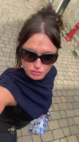A woman with dark hair and sunglasses is standing outside and holding a six-pack of Nivea deodorant spray. She is wearing a dark blue sweater draped over a black tank top and black leggings. A set of keys hangs from a clip on her left hip. The background shows a cobblestone path and a stone building. She turns the product to show the label and the camera.