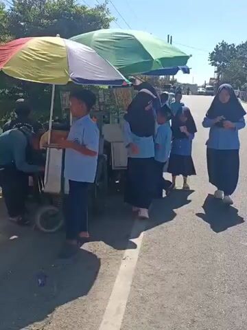The video shows a group of children, seemingly students based on their uniforms, gathered around a street vendor's cart. The cart is covered by a multicolored umbrella, with green and yellow panels visible. It appears to be a food or snack cart, as one child is holding a tray with what looks like food. The children are wearing light blue shirts and dark skirts or pants, and several of the girls are wearing hijabs. The scene is outdoors, on a paved road with a white line marking the edge. The sun is shining brightly, casting shadows. In the background, there are trees and a few buildings. The children are interacting with the vendor and with each other, some appearing to be waiting in line or making a purchase. The overall impression is a mundane street scene of children buying snacks.