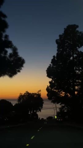 The video shows a view of a road leading downhill towards the ocean at sunset. The sky is a gradient of blue at the top, transitioning to orange near the horizon. Silhouetted trees frame the road on both sides. Cars are parked along the road and at least one is driving. Yellow lines mark the lanes. Utility poles are visible, and a street light is illuminated. The ocean horizon is a bright orange.