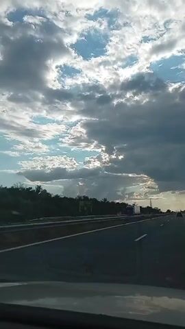 The video shows a view from inside a vehicle traveling on a highway. The road is paved, with white lines marking the lanes. There is a line of trees or bushes along the side of the road. In the distance, there are power lines and what appears to be a truck. The sky is filled with dramatic clouds, some dark and heavy, others bright with sunlight breaking through. The sun is low in the sky, casting long shadows. The overall impression is of a journey on a highway under a changeable sky.