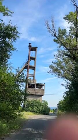 The video shows a tall, concrete mining tower under a bright blue sky with scattered clouds. The tower is partially obscured by green trees and bushes in the foreground. It appears to be an abandoned structure, with some visible rust and deterioration. The camera slowly zooms in on the tower, revealing more details of its construction and the surrounding landscape. The tower has multiple levels and an inclined structure leading up to its top. Power lines are visible stretching across the frame in the middle distance. The scene is outdoors on a sunny day.