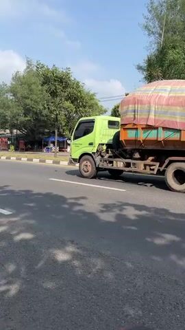 A light green dump truck with a tan and red striped tarp covering its load drives down a paved road. The truck has a chrome bumper and a dark colored cab. The tarp is bulky and appears to be made of a heavy duty material. The truck is in the right lane of a two-lane road, with white and yellow lane markings. The road appears to be in a rural or suburban area, with trees and some buildings visible in the background. The sky is blue with some clouds. The video appears to be filmed from another vehicle on the road.
