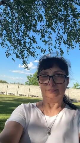 This is a selfie-style video featuring a middle-aged woman with dark hair, bangs, and glasses. She is wearing a white t-shirt and a silver necklace with a pendant. The woman is outdoors, with a birch tree and its leaves in the foreground, partially obscuring the view. Behind her, there's a well-maintained grassy lawn and a light-colored, segmented fence. The sky is a clear blue with a few scattered white clouds, suggesting a pleasant day. The sunlight indicates it is likely daytime, possibly late morning or afternoon. The woman appears calm and is looking directly at the camera with a slight smile. The overall scene conveys a relaxed, outdoor setting, likely a backyard in Hnarovske, Ukraine. There is no visible text in the video.