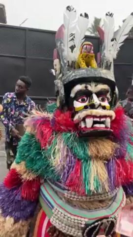 The video shows a costumed figure in what appears to be a public celebration. The figure wears a large, colorful feathered costume and an elaborate mask. The mask is painted with white, black, yellow, and red colors, and has protruding teeth. On top of the mask is a helmet-like structure adorned with white feathers and a small yellow animal figure. The figure is holding and waving a bill. Other people are visible in the background, some in ordinary clothes, and some in the red shirts. In the background, a speaker. The video seems to be taken outdoors on a cloudy day.