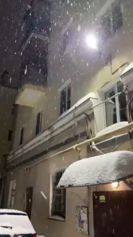 The video shows a nighttime scene during a heavy snowfall in a courtyard. A pale yellow building with windows and white trim stands to the right of the frame, partially illuminated by a bright streetlight above.  Snow has accumulated on a small awning and various horizontal surfaces. To the left, a darker building can be partially seen. Snowflakes are visibly falling, creating a dense white shower in the illuminated area. A car is partially visible in the lower left corner. The camera angle is looking upwards at the buildings.