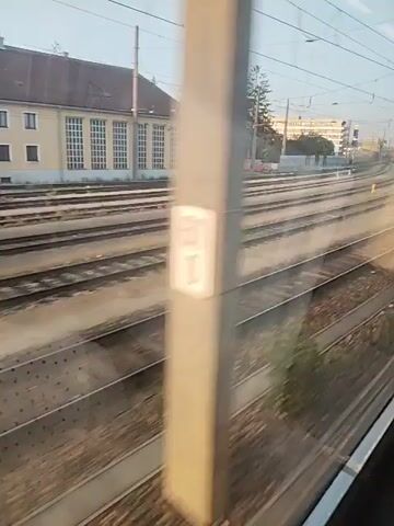 The video shows a train moving through a railway yard in Sankt Pölten, Austria. The scene is dominated by multiple sets of train tracks stretching into the distance, with a low-rise building that appears to be a station or administrative building on the left. Several freight trains are visible on the tracks, some of which are covered in graffiti. The time of day appears to be morning or late afternoon, given the low angle of the sun and the warm lighting. The weather seems clear and calm. The camera is positioned inside a moving train, looking out the window, giving a sense of motion and passing scenery. The building on the left has a light-colored facade with multiple windows and a pitched roof. There are overhead power lines for the electric trains, typical of European railways. The overall impression is that of a functional industrial or transport hub. No people are clearly visible. There is text on the side of one of the freight cars, "STBR EFT".