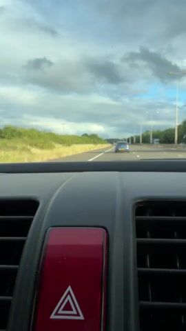 The media file is a first-person perspective shot from inside a car, looking forward through the windshield. The primary focus in the foreground is the car's dashboard, specifically the hazard light button, which is red with a white triangular symbol. To the left and right of the button are air vents.

The view through the windshield reveals a highway with a blue car in the distance. The road is bordered by a grassy verge and lush green trees and bushes. In the background, the sky is filled with dramatic, cloudy formations, suggesting a dynamic weather system, but the sunlight breaking through indicates it's likely late afternoon or early evening. The overall setting appears to be a rural or semi-rural area, consistent with the provided location context of the Malvern Hills in the United Kingdom. There is no visible text in the media file itself, although there are timestamps indicating the video is only 4 seconds long. The activity is travel on a highway.