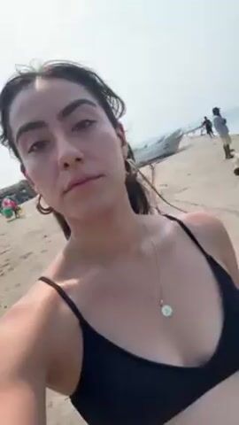 A young woman in a black bikini takes a selfie on a beach. She has dark hair pulled back, hoop earrings, and a necklace.  The beach has tan sand and other people walking in the background. A small boat sits on the sand behind her. The ocean is visible with waves coming to shore. The sky is overcast.