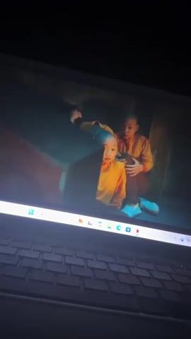 A laptop is displayed in a dimly lit room, with its screen showing a video. The video features a dynamic martial arts fight scene set within an ornate indoor environment, characterized by traditional Eastern architecture and a light blue/teal colored floor. A central figure, dressed in a flowing orange robe, appears to be a martial arts master or monk. This individual is shown engaging in combat with multiple adversaries clad in dark, possibly armored, costumes. Swords are visible as weapons during the action. Subtitles on the screen indicate specific audio cues: "[suspenseful music playing]" and subsequently "[grunting]". The laptop's keyboard is partially visible in the foreground. The illuminated taskbar at the bottom of the screen displays various application icons, including the Windows logo, search, several web browsers, Microsoft Office applications (Word, Excel, PowerPoint, OneNote, Outlook), Spotify, and Netflix, alongside system icons for Wi-Fi, volume, and battery.
