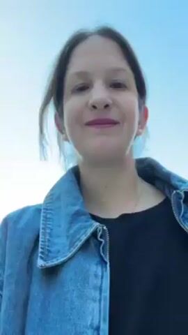 A woman is taking a selfie video, rotating to capture the view of a small harbor. The woman has shoulder-length brown hair, pink lipstick, and is wearing a blue jean jacket over a black shirt.  She is standing on a concrete walkway with a rope railing alongside the water. In the background, there is a rocky coastline with some buildings on top.  There are a few people walking on the rocks by the water's edge.  The water in the harbor is calm and blue. The sky is clear. The woman makes a kissing face at the camera.