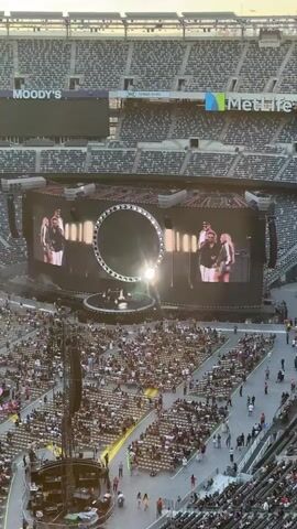 The video shows a concert from a high-angle, elevated perspective looking down at a large stadium. In the center of the frame is a massive stage set up with multiple large screens. The central screen displays animated graphics and lyrics. The screens show two people, likely performers, on either side of a large circular graphic that displays words like "RISE" and "WHAT" sequentially. The stage itself appears to be circular with smaller circular elements and lighting. Below the stage, rows upon rows of stadium seating are filled with a large audience. Some people are standing and walking in the aisles. The stadium has tiered seating and large advertising boards are visible at the top of the stadium structure, including "MOODY'S" and "MetLife". The lighting suggests it is either late afternoon or early evening. There are also large speaker towers and lighting rigs visible in the foreground and around the stage. The overall scene is a massive outdoor concert venue.