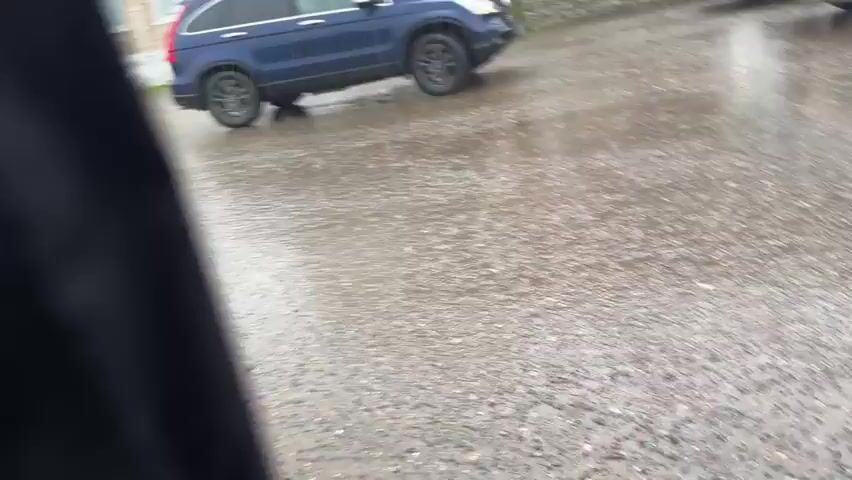 The video captures a static, eye-level view of a wet street scene on a rainy day. Heavy rainfall is evident, with puddles forming on the asphalt and rain streaks visible in the air. The ground is thoroughly wet and reflective.

Three cars are prominent in the foreground and midground. On the left, a dark blue SUV (resembling a Honda CR-V) is parked or stationary, facing slightly right. To its right, further back, a white sedan is parked. On the far right, a light grey or silver SUV (a modern crossover model) is parked, facing left. All vehicles appear wet from the rain.

In the background, a multi-story brick building with several windows lines the street. The ground floor features several storefronts. Clearly visible above one entrance is a red sign with white Cyrillic text reading "ГОСАПТЕКА" (GOSAPTEKA), indicating a State Pharmacy. Below this, another sign reads "АССОРТИ" (ASSORTI). Further to the right, a partially obscured blue sign has "ПРОДМАГИ" (PRODMAGI) written on it, suggesting a grocery or food store.

One individual, dressed in dark clothing, walks up a few steps towards the pharmacy entrance. Another person is briefly visible inside the pharmacy doorway. The overcast lighting suggests daytime, but the heavy rain creates a somewhat subdued atmosphere. The scene depicts a typical urban street during a downpour, with everyday activities continuing despite the weather.
