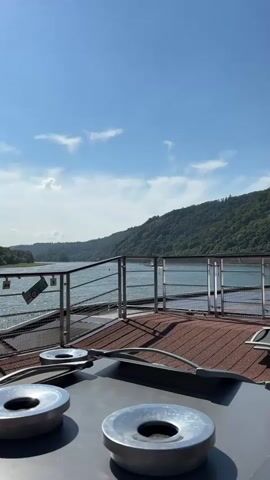 A vertical, handheld shot looking out from the deck of a boat or ship. In the foreground, two round, metallic objects with circular openings are visible, possibly ashtrays or part of a table. Behind these, a deck area with reddish-brown textured flooring and several rows of empty lounge chairs with metallic frames and dark seats are visible. A metal railing separates the deck from the water. Beyond the railing, a wide body of water stretches into the distance, lined by lush green, tree-covered hills. The sky is a clear blue with a few scattered white clouds. A small flag with a green, red, and white horizontal striped pattern and a star is attached to the railing on the left side of the frame. A white building with a dark window and two white dome-like structures on its roof is situated on the far side of the water, on the edge of the hillside. The overall scene suggests a sunny day with calm weather on a river or lake.