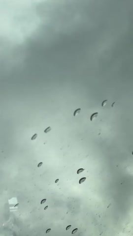 The video shows raindrops on a windshield against a cloudy sky. The camera is static and focuses on the water droplets, some of which are clustered together while others are isolated. The background is out of focus, creating a bokeh effect. The lighting is dim, suggesting an overcast or rainy day.
