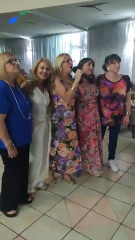 The video shows a group of five women standing in a line and singing, holding a microphone. They are in an indoor setting, possibly a restaurant or hall, with a patterned tiled floor and light-colored walls. The women are dressed in colorful attire, with one in a blue top and black pants, another in a white jumpsuit, two in floral dresses, and the last in a brightly colored patterned top and dark pants. The woman in the middle is holding a microphone and appears to be leading the singing. The other women are looking towards the microphone and singing along. The lighting suggests it is daytime. The mood seems festive and celebratory. There is no visible text in the video.