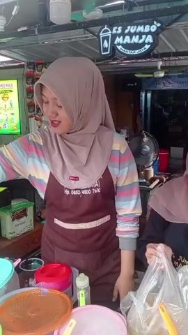 A woman wearing a headscarf and an apron is standing behind a food stall. She is holding a hand over a green container. To her right, another woman in a headscarf is holding a plastic bag. In the foreground, there are several large plastic containers with colorful lids, and stacks of clear plastic cups. The stall has a sign that reads "ES JUMBO MANJA MANTAP JAYA". There is also a menu board visible on the left side of the stall. The environment appears to be outdoors, possibly a street food vendor setting.