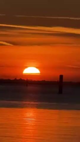 The video shows a vibrant orange sunset over the water. The sun is positioned low on the horizon, appearing as a semi-circle due to its descent.  The sky is painted in various shades of orange, with thin streaks of clouds. The water reflects the sun's color, creating a shimmering effect. Several wooden posts are visible, punctuating the water near the horizon.