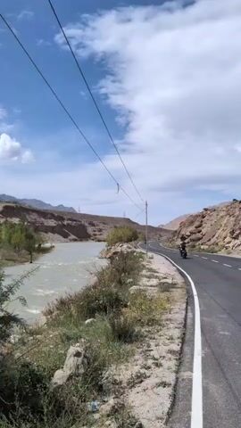 A paved road curves alongside a river. The river is wide and appears to be flowing swiftly, with murky water. On the left bank, there are hills with sparse vegetation. On the right side of the road, there are rocky, barren hills. There are utility poles and wires running parallel to the road. Two motorcyclists are visible on the road, further ahead. The sky is blue with scattered clouds. The overall impression is a remote, mountainous landscape.