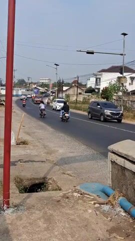A vertical video captures a road scene with traffic under a clear sky. On the left, a red pole stands near a sidewalk with a drain grate and blue pipes. The road curves to the right. Several motorcycles and a few cars, including a white van and a dark SUV, are traveling on the road. A red truck with a black roof and front is also visible, as is a white building with a black roof in the background, surrounded by trees and other houses. Solar-powered streetlights are present along the road.