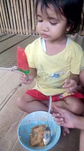 A young child is seated on a woven mat, holding a red and green popsicle in their left hand. They wear a yellow t-shirt with a graphic print and red shorts. The child is looking down at the popsicle, their eyes closed. To the right of the child is a blue bowl containing a light brown, mushy substance, with a spoon resting in it. Another person's hands are visible in the upper right corner, holding the spoon as if feeding the child from the bowl. The child appears to be in the process of eating the popsicle, with a small bite taken out of it. The setting is indoors, with a cardboard surface beneath the mat and bamboo structures to the left. The lighting suggests it could be daytime, possibly with natural light. The overall mood seems relaxed, with the child engrossed in their treat.