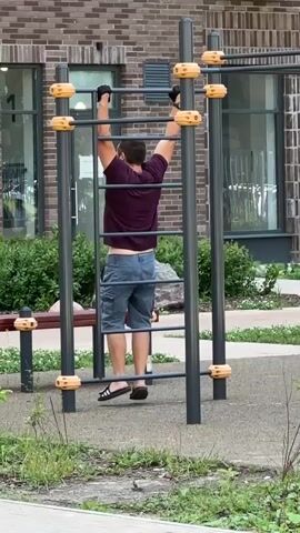A man is exercising on a playground pull-up bar. He is wearing a dark red t-shirt, blue shorts, and black gloves. He grips the bar with both hands and performs leg raises, lifting his legs towards the bar. The playground equipment is gray with yellow grips. In the background, there is a brick building with windows and green bushes. A bench is also visible behind the pull-up bar. The ground is covered with gravel and some grass.