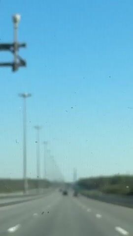 The view from a moving vehicle on a multi-lane highway with streetlights shows a tall, pointed building in the distance. The sky is clear and blue. The road appears to be mostly empty. The car moves forward, with the building visible between the streetlights.