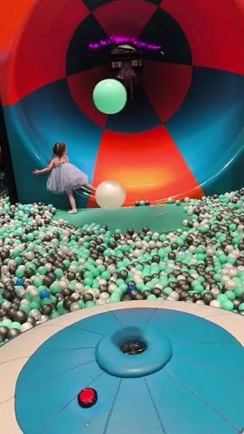 A young girl wearing a light blue tutu stands in a large ball pit, with her back to the camera. She is positioned on a raised platform that appears to be the entrance to a large, colorful tube slide. Two large, inflatable balls, one light green and one white, are suspended in the air in front of her, slightly above the ball pit. The slide itself is a wide, curved tube with sections of red, blue, and teal. In the distance, further up the slide, another young child is visible, also standing. The ball pit is filled with numerous small, round balls in various shades of green, grey, and white. In the foreground, a large, round, blue structure with a red button is visible. The overall environment appears to be an indoor play area.