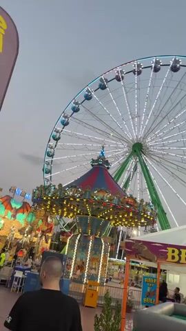 The video shows a daytime scene at a carnival or amusement park. In the foreground, a person wearing a black t-shirt with white lettering is walking away from the camera. To their left, another person in a white shirt is partially visible. In the background, a large Ferris wheel with white lights is prominent against a light gray sky. Below the Ferris wheel, a colorful carousel with blue, red, and gold accents is in operation, adorned with numerous lights. To the left of the carousel, a brightly lit attraction with a castle-like facade and colorful banners is visible. Fencing separates the viewing area from the rides. Further to the right, a sign with the words "RIDE THE BULL" is visible, suggesting a bull riding attraction. The ground is a reddish-pink color. The overall atmosphere appears to be lively and festive.