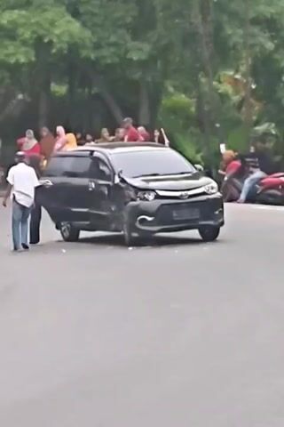 The video captures a street scene with a damaged black van parked on the right side of the road. The front of the van is visibly crumpled, suggesting an accident. Several people are gathered around the vehicle, some appearing to be inspecting the damage or conversing. On the left side of the road, more people are standing, and a few motorcycles are parked. The background features lush green trees and foliage, indicating an outdoor, possibly suburban or park-like setting. The ground is asphalt, typical of a road surface. The lighting suggests it is daytime. The overall atmosphere seems to be one of a minor incident with onlookers.