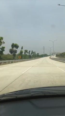 A POV shot from inside a vehicle driving on an empty highway. The road is made of concrete and has multiple lanes. The sky is overcast and hazy. On the left side of the road, there is a guardrail, trees, and power lines. On the right side, there are streetlights and highway signs. The vehicle appears to be moving at a moderate speed. No other vehicles or people are visible in the immediate vicinity.