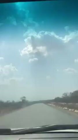 The video depicts a view from inside a vehicle driving along a paved road. The sky is bright blue with scattered white clouds. The road stretches ahead, flanked by dry, grassy terrain and sparse vegetation. The windshield has a blue tint and shows reflections.  There are no other vehicles visible, but what appears to be a motorcycle is approaching in the distance on the right side of the road. A sticker of "Elizabeth Masiyiwa" is affixed to the front windshield. The overall impression is one of a sunny day on a relatively empty road.