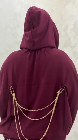 This video shows the back of a person wearing a burgundy hooded jacket. The jacket has gold chain detailing attached to the back, with safety pins holding the chains in place on either side of the jacket. The chains hang down the back of the jacket. The background appears to be a wall with a textured finish.