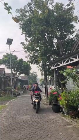 A person is riding a motorcycle down a paved street. The person is wearing a dark blue shirt and has two young children with them. One child is sitting in front of the person, and the other child is sitting behind them. The children are wearing red shirts. The street is lined with houses and trees. There are clothes hanging on a clothesline in front of one of the houses. The sky is cloudy.