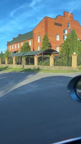 The media file captures a brick building with the Cyrillic text "ДІМ МОЛИТВИ" (House of Prayer) visible on its facade. The building has a dark green roof and several arched windows. A covered entrance with a metal awning leads to the main doors, accessible by a set of stairs with metal railings. The building is surrounded by a fence with decorative metalwork and brick pillars. Green trees and manicured bushes are planted around the property.

The scene appears to be a street view, as indicated by the asphalt road in the foreground and a car's side mirror reflecting the scene. The time of day is suggested by the long shadows cast on the road and the bright, clear blue sky with a few wispy clouds. The lighting suggests it's either early morning or late afternoon. The weather appears to be fair.

The overall impression is one of a serene and well-maintained religious or community building. No people are visible in the footage. The location is identified as Dovhe, Ukraine.