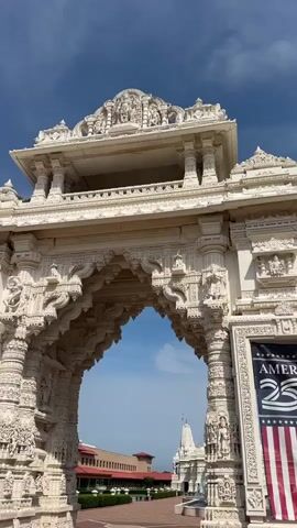 The video captures a close-up view of an intricately carved white stone archway leading to a larger complex. The archway features detailed sculptures and patterns, with columns on either side adorned with figures. Through the arch, a white temple with multiple spires is visible in the distance, set against a blue sky with some clouds. To the left of the temple, a long, reddish-brown building with a red roof is partially visible. The ground in front of the archway is paved with reddish-brown bricks laid in a pattern. A banner with American flag elements and the text "AMERI" is attached to the right side of the archway. The camera appears to be stationary or moving very slowly, focusing on the ornate archway and the scene beyond.