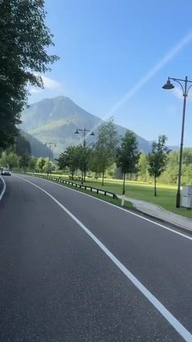 The video shows a road with a white line down the middle. There are trees on the left side of the road, and a mountain in the background. There are also streetlights along the road. A white van with bicycles on the back drives past. There is a grassy area on the right side of the road with more trees and some buildings in the distance. The sky is blue with some clouds.