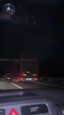 The video shows a car driving on a dark highway at night. The camera is positioned inside the car, looking through the windshield. Initially, the car is passing a large truck on the left lane. As the car moves forward, the truck is left behind and the road ahead is illuminated by the car's headlights and the taillights of other vehicles in the distance. The road appears to be well-maintained with clear lane markings. The inside of the car is visible, including the dashboard and the hazard light button. Overall, the environment suggests a typical nighttime highway drive with limited visibility.