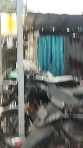 The video pans across a cluttered outdoor area with several motorcycles parked under a covered structure. In the foreground, a portion of a motorcycle's front tire is visible. The camera then moves to reveal a collection of dirt bikes, some of which appear to be older models, parked together. One dirt bike in the center of the frame is green and white with racing decals and the number 77 prominently displayed. The background shows a corrugated metal roof, some wooden structures, and large yellow water tanks. The overall environment appears to be a storage or workshop area for motorcycles, with a focus on dirt bikes.