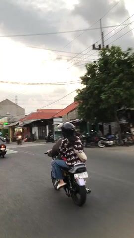 The video shows a street scene with several motorcycles. In the foreground, a motorcycle with two riders is moving away from the camera. The rider in front is wearing a helmet and a striped shirt, and the passenger behind is wearing a helmet and a black and white patterned shirt. In the background, other motorcycles are visible on the street. The environment appears to be a town or city with buildings lining the street and overhead power lines. The sky is cloudy, suggesting it may be dusk or overcast. The overall impression is of everyday street life with motorcycle transportation.