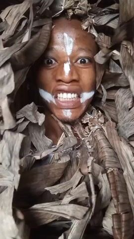 A close-up shot focuses on a person covered in dried banana leaves. The person has brown skin, wide eyes, and white paint on their forehead and around their mouth. They appear to be indoors. The person is seen blowing air out of their mouth and moving their face.