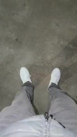 A person wearing white sneakers and gray jeans is walking on a gray surface, likely a sidewalk. The camera is positioned at a high angle, looking down at the person's feet and legs. The person walks past a metal fence.