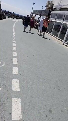 The media file shows a sunny day in Teguise, Spain, with clear blue skies and bright sunshine. The scene is an outdoor path or walkway, likely a promenade or street, with a gray asphalt surface marked with white dashed lines. In the background, there is a white building with a rooftop terrace. The building has large windows or glass storefronts displaying merchandise, suggesting a commercial area such as shops or restaurants. The text "Blue Note" is visible on a sign above the building's entrance.

Several people are walking along the path. On the left, a group of three individuals are walking away from the camera. The first person is a man wearing a dark t-shirt and shorts. Next to him is a woman in a pink dress. Behind them, partially obscured, is a person in a white shirt and shorts. Further to the right, a person in a colorful, patterned short-sleeved shirt and black shorts is walking towards the camera, holding their hands up to their head as if shielding their eyes from the sun or adjusting their hair. Another person in a white shirt and shorts walks beside them. Further in the distance on the left, a group of people appear to be cycling or riding bicycles.

The overall atmosphere suggests a warm, pleasant day, typical of a tourist destination. The people appear to be casually dressed, likely enjoying their time outdoors. The presence of shops and people strolling implies a public space, possibly near a beach or a tourist attraction.