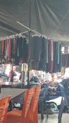 The video shows a market stall with clothes hanging on display. There are several people in the background, some of whom appear to be customers or vendors. The stall is covered by a black tarp, and there are chairs in the foreground. The lighting suggests it is daytime.