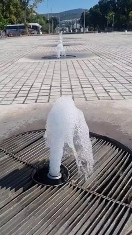 The video shows a series of small, powerful water fountains spraying upwards from circular grates embedded in a tiled plaza. The closest fountain is in the foreground, with a tall column of water that splatters outwards. In the background, several more fountains are visible in a line, receding into the distance. The plaza is paved with light-colored square tiles. Beyond the plaza, there are trees and buildings, and a clear blue sky. A bus and some people are visible in the far background on the left side of the frame. The overall impression is an outdoor public space with decorative water features.
