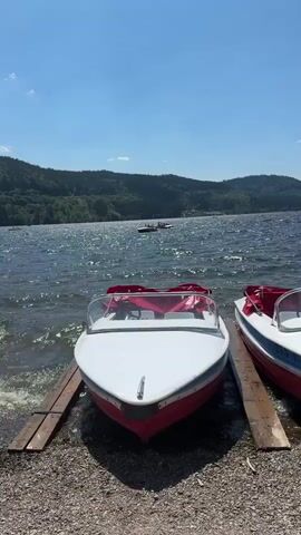 A video shows a close-up view of two small boats docked on a gravelly shore. The boats are white with red accents and have red seats. The water is choppy and reflects the sunlight. In the background, there is a green, forested hill under a clear blue sky. There are other people on the water in a distant boat or raft.
