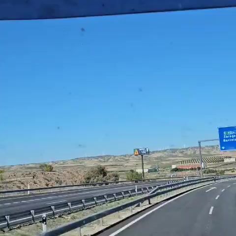 The video shows a view from inside a vehicle moving along a highway on a sunny day. The sky is clear and blue. The landscape includes rolling hills and fields. Wind turbines are visible in the distance. The highway has a barrier on the left side. A sign displays the price of "Gasóleo 287" as "1.482". The vehicle continues driving forward along the highway. The sign briefly changes to "8.888".
