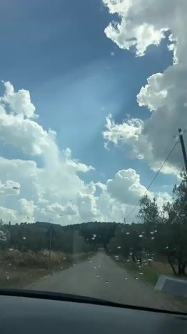 The video is filmed from the inside of a car looking through the windshield at a rural road. The road is paved and appears to be a single lane. On either side of the road are olive trees and dry grass. The sky is blue with large, fluffy white clouds. Sunbeams are visible breaking through the clouds. There are water droplets on the windshield. A signpost is visible on the left side of the road. The video is shaky, indicating the car is in motion.