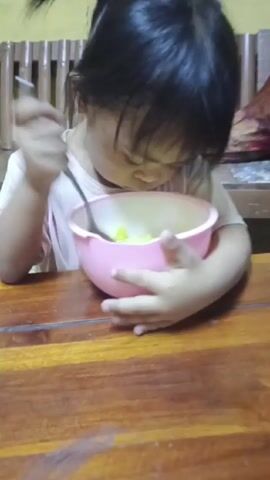 A young child with dark hair in a ponytail is sitting at a wooden table, eating from a pink bowl with a spoon. The child holds the bowl with their left hand and brings the spoon to their mouth with their right. The bowl appears to contain something yellow with green chunks. The background shows a wooden bench.