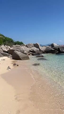 The video shows a tranquil beach scene with a clear blue sky and calm, turquoise water lapping gently against the shore. Large, smooth rocks are scattered along the coastline, with some submerged in the water. The sand is light-colored and appears fine-grained. Lush green vegetation can be seen on a hillside in the background to the left. The water is so transparent that the sandy bottom and submerged rocks are clearly visible. The overall atmosphere is peaceful and serene, typical of a tropical island or secluded cove. There are no people or discernible objects other than the natural elements of the beach.